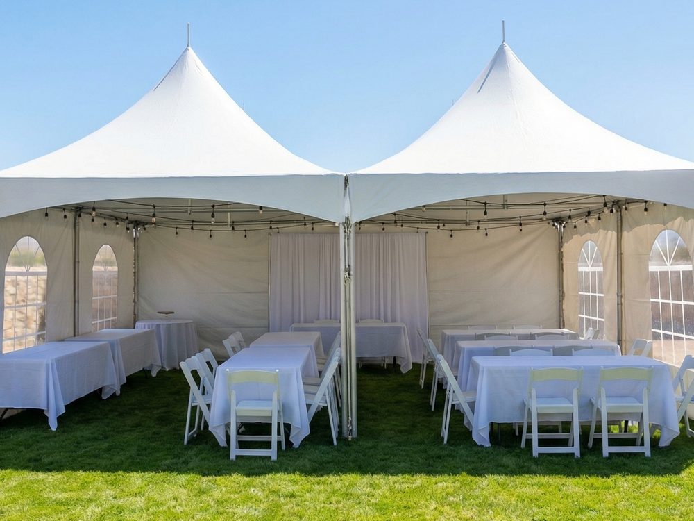 Two high-peak tents joined side by side on a lawn, sidewalls up, banquet tables and folding chairs inside.